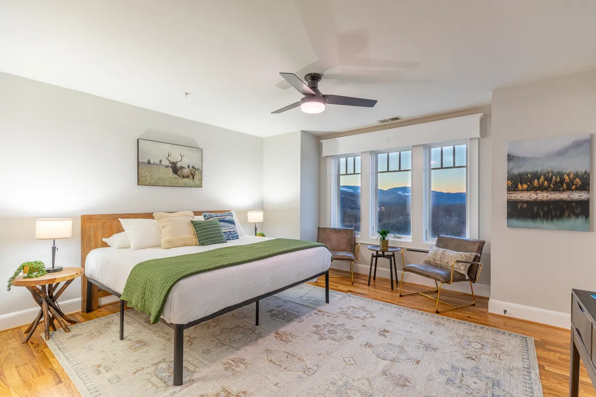 A tranquil queen bedroom with a queen-sized bed, large windows letting in natural light, tasteful artwork, and stylish armchairs flanking the bed for added comfort. Nightstands with lamps offer functional elegance.