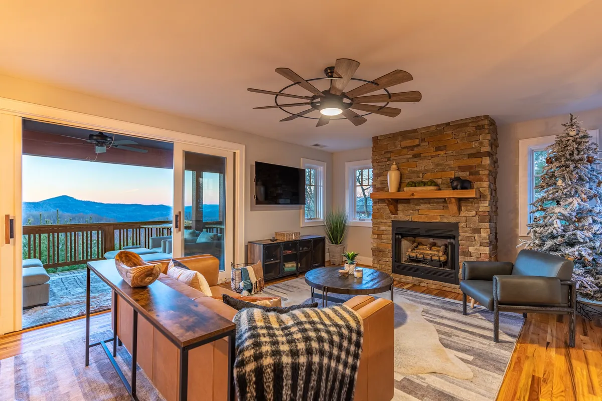 The image shows a warm, inviting living area featuring a stone fireplace, plush seating arrangements, and expansive windows framing stunning mountain views. A decorative tree adds a touch of charm, while natural light pours in, illuminating the cozy space and highlighting its welcoming ambiance.