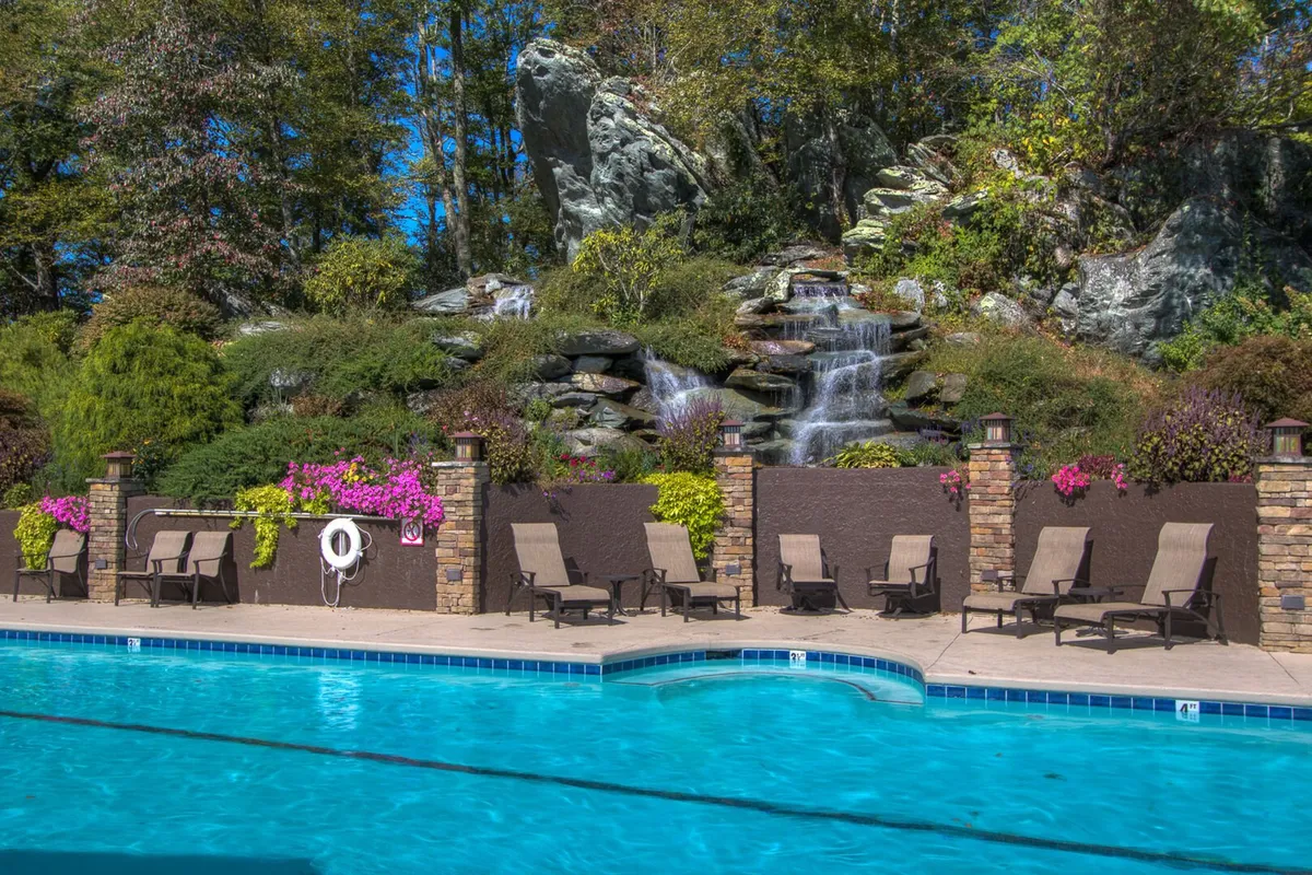 A beautifully landscaped outdoor swimming pool with a cascading waterfall, surrounded by lounge chairs and vibrant flowering plants. The setting is tranquil, inviting guests to swim or unwind by the pool while enjoying scenic natural views.