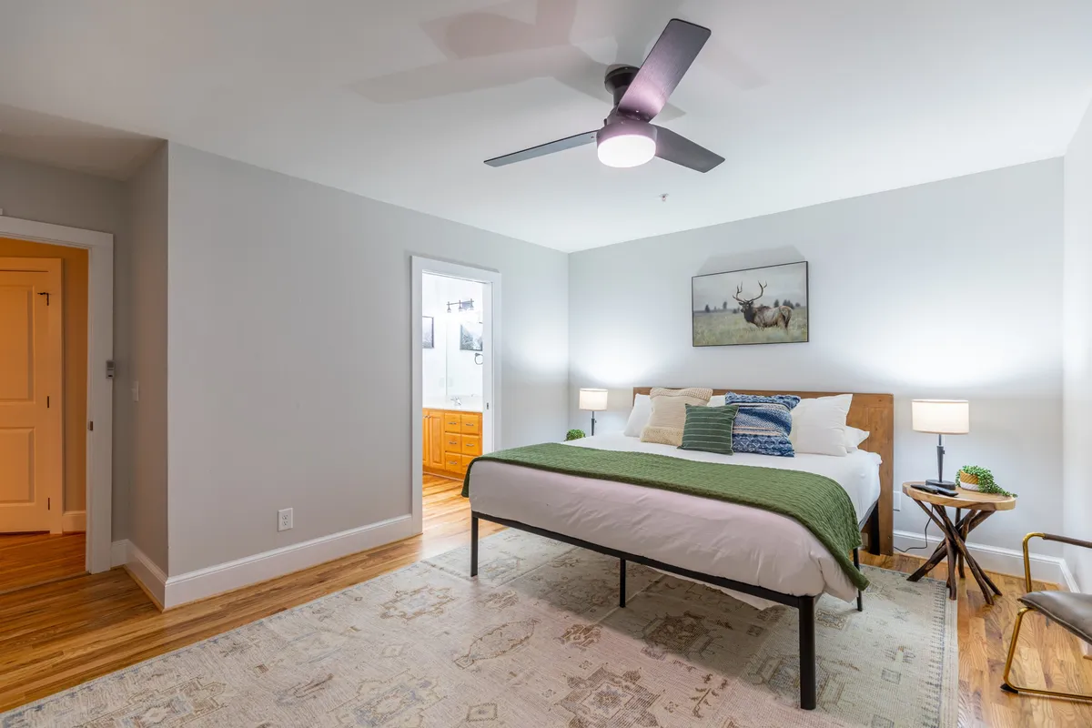 A tranquil bedroom featuring a large bed with a green throw blanket, nightstands with softly glowing lamps, and tasteful wall art of a deer. Natural light streams through the window, enhancing the room's peaceful ambiance.