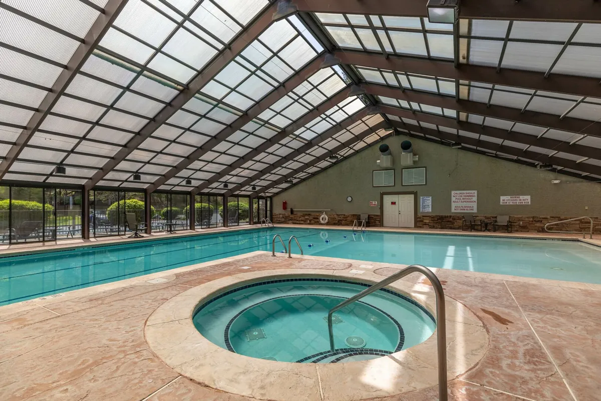 A bright, spacious indoor swimming pool area featuring a sparkling pool and a hot tub under a large glass roof. There are ample seating options around the pool, creating a welcoming space for both relaxation and social gatherings regardless of the season.