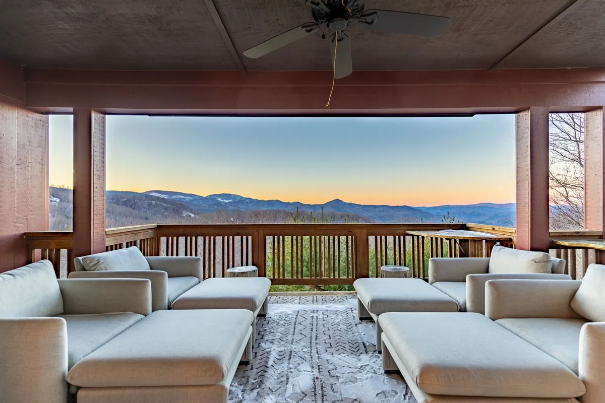 An inviting outdoor seating area on a covered deck, featuring plush lounge chairs atop a textured rug, all oriented toward a breathtaking view of the Blue Ridge Mountains. The space is under cover, suggesting comfort in all seasons and a seamless transition between indoor coziness and mountain vistas.