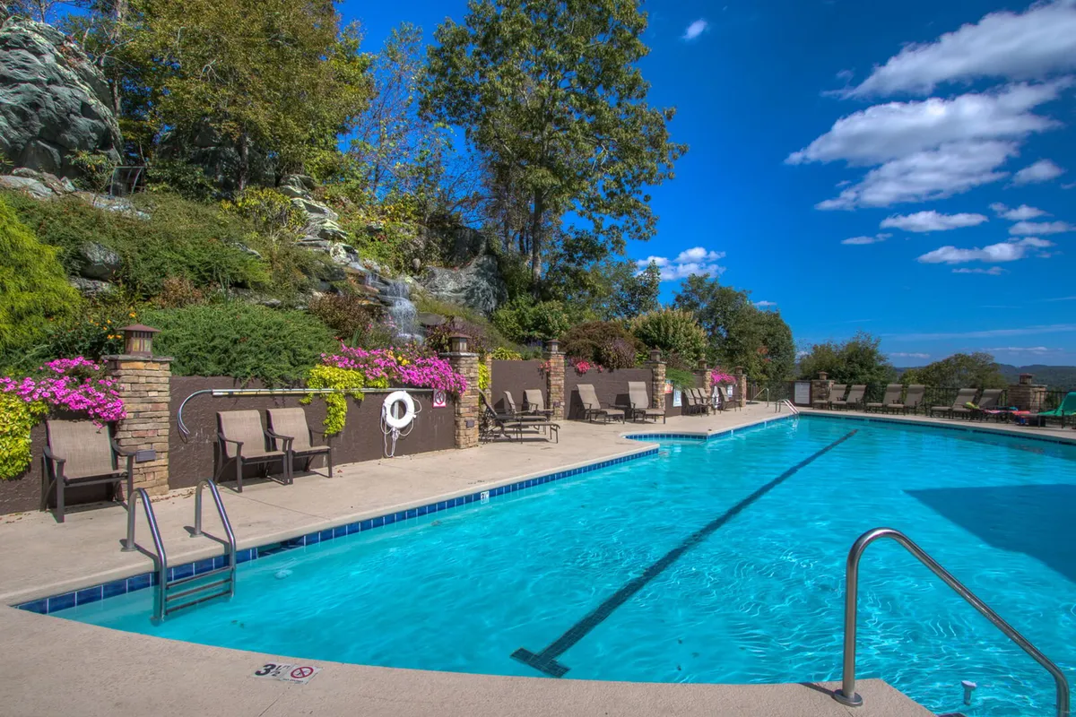 A sunlit outdoor swimming pool surrounded by a natural, rocky landscape, vibrant flower beds, and comfortable lounge chairs. The area gives off a tranquil yet lively atmosphere, perfect for socializing or relaxing by the water. The pool area is set against a backdrop of lush greenery, highlighting the connection to the outdoors and resort-style amenities.
