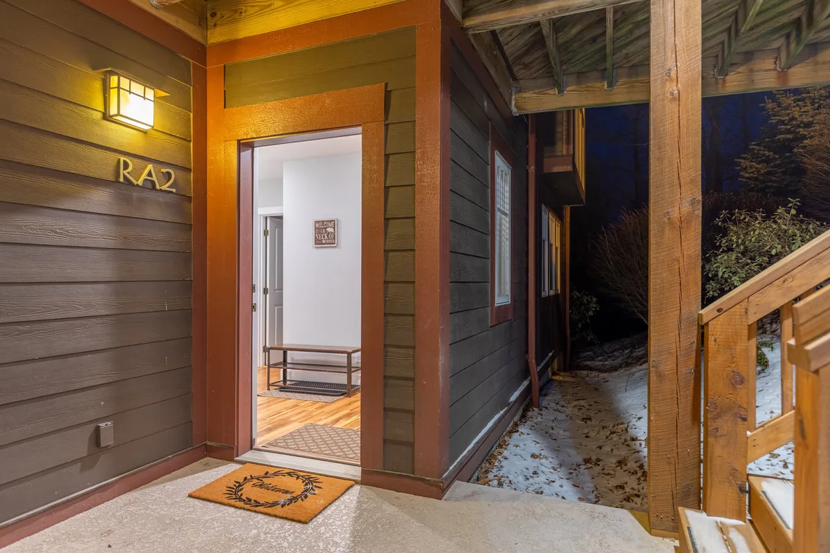 The welcoming front entrance of the property, featuring a charming wooden front door, a welcome mat, and inviting outdoor lighting. Wooden steps lead up to the entry, surrounded by natural elements that evoke the mountain setting.