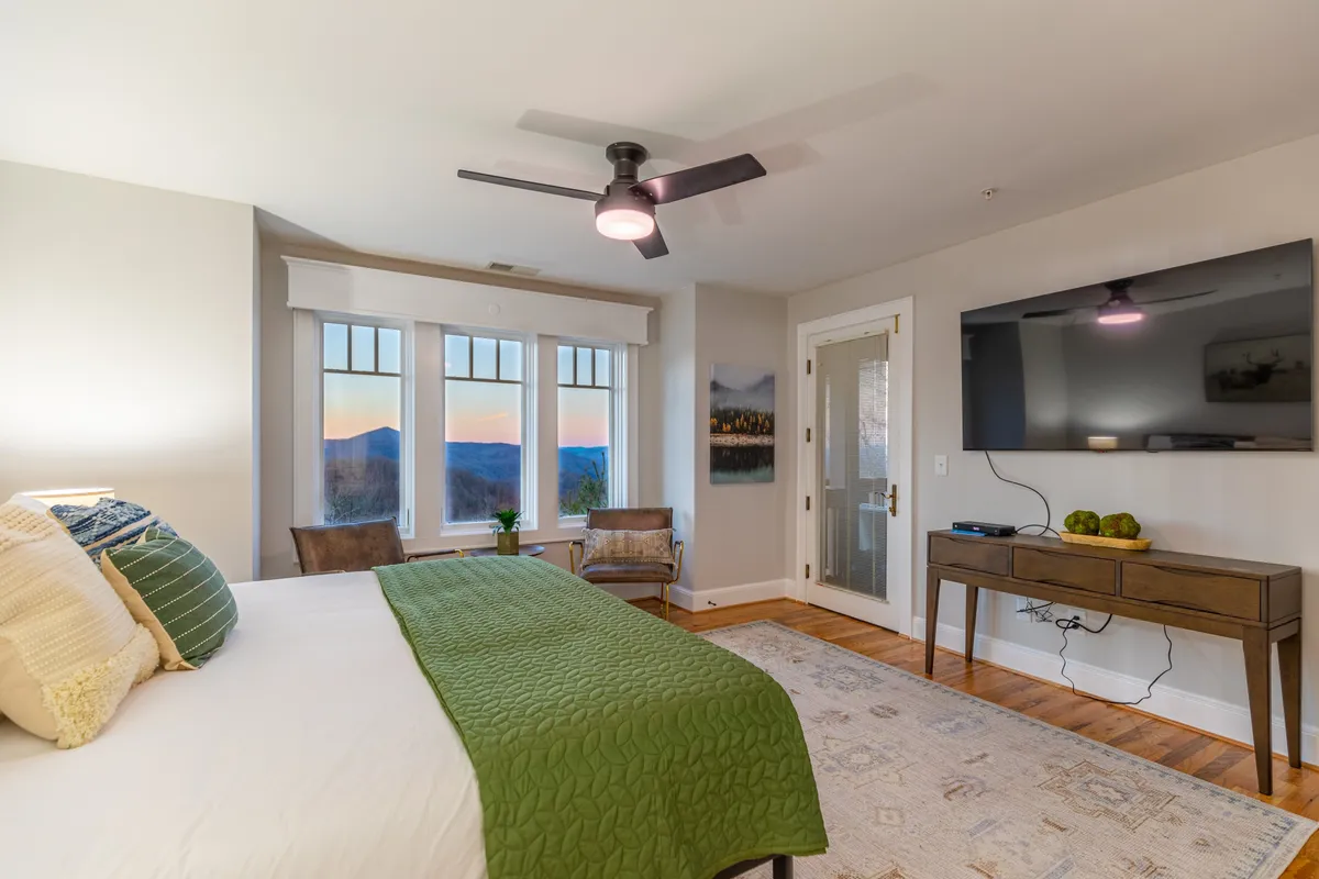 A spacious bedroom featuring a comfortable bed with a green blanket, large windows that frame mountain views, a sleek modern TV setup opposite the bed, and decorative artwork providing a cozy ambiance.