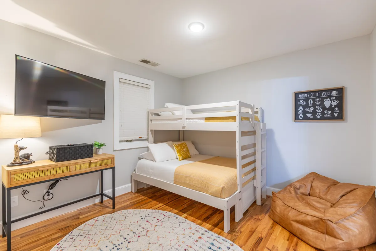 A playful, inviting bunk room featuring a twin-over-full bunk bed, a television mounted on the wall, colorful wall art, a bean bag chair, and a soft area rug that encourages relaxation and fun.